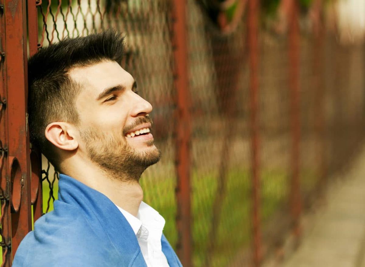 A young man with short dark hair and a beard, smiling warmly while leaning his head against a rusted metal fence in soft sunlight