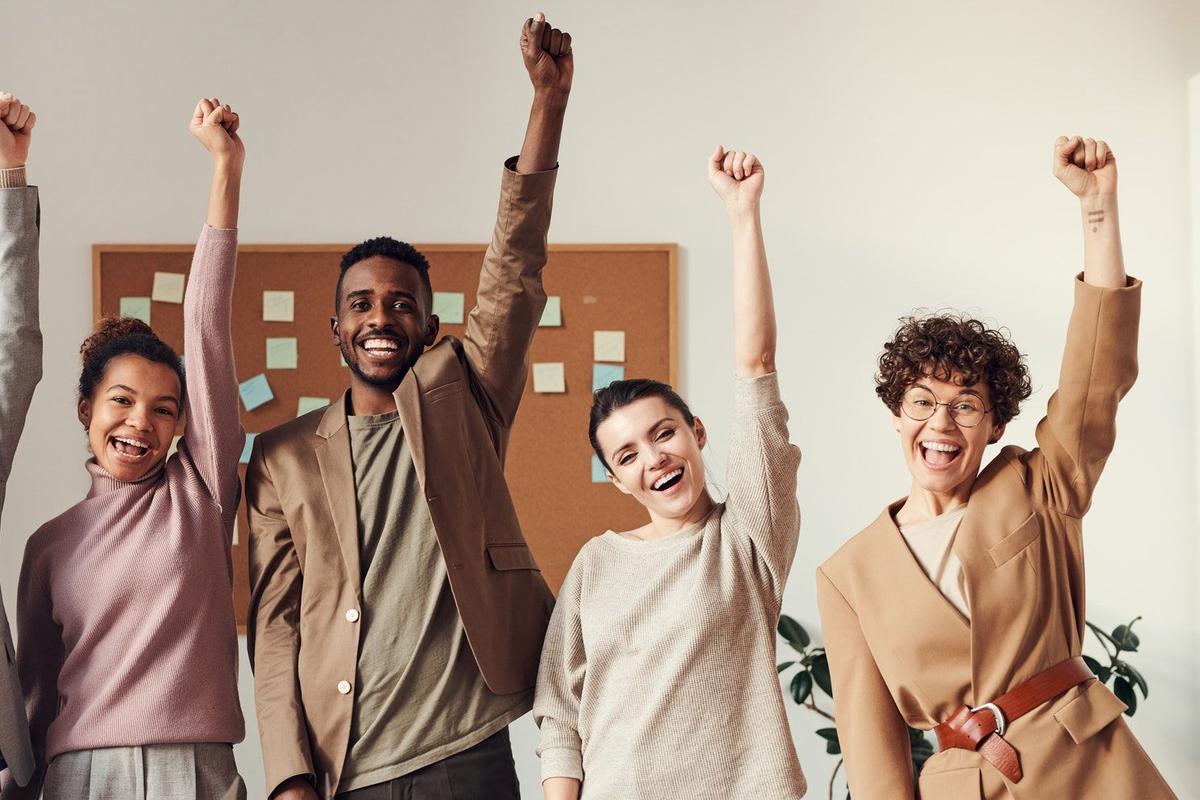Diverse group of coworkers celebrating success in an office, smiling and raising their fists in the air during a team meeting, symbolizing motivation, teamwork, and achievement.