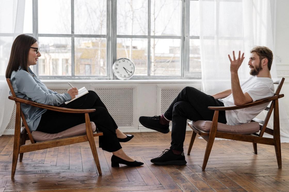 A female therapist sitting in a wooden armchair takes notes while listening to a bearded male client who is gesturing with his hand during a therapy session in a bright, modern office.