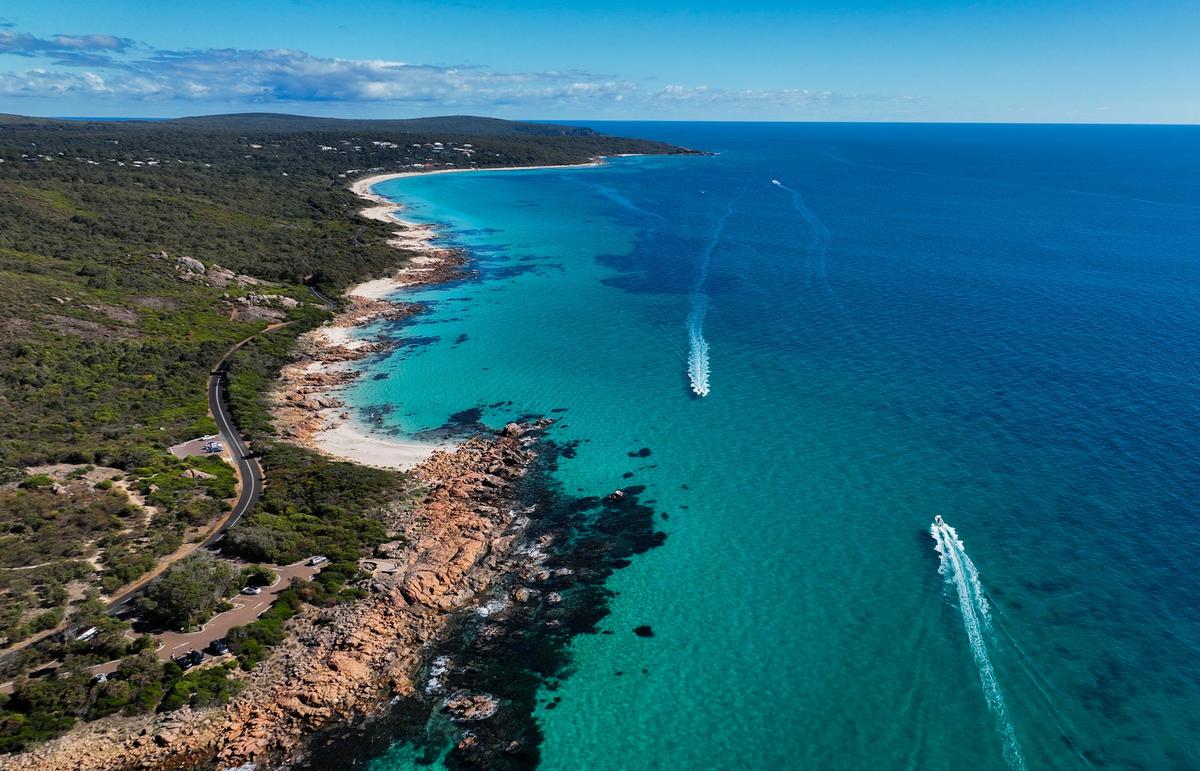 An aerial, high-angle view of a turquoise coastline with two boats leaving white wakes in the deep blue ocean.