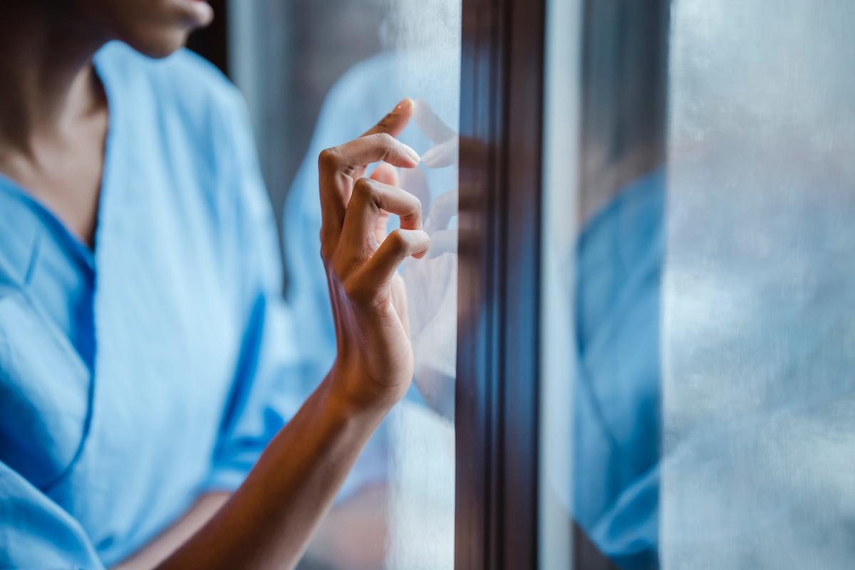Person in blue clothing gently touching a window while looking outside, representing reflection and support during bipolar disorder treatment.