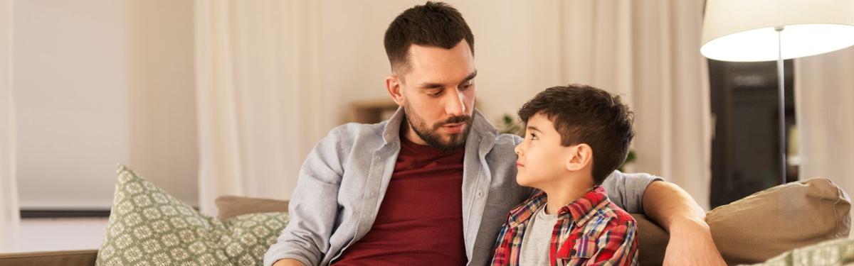 Father sitting on a couch with his young son, making eye contact during a serious conversation about difficult topics at home.