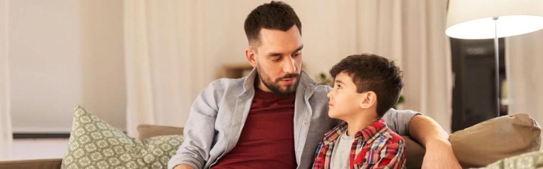 Father sitting on a couch with his young son, making eye contact during a serious conversation about difficult topics at home.