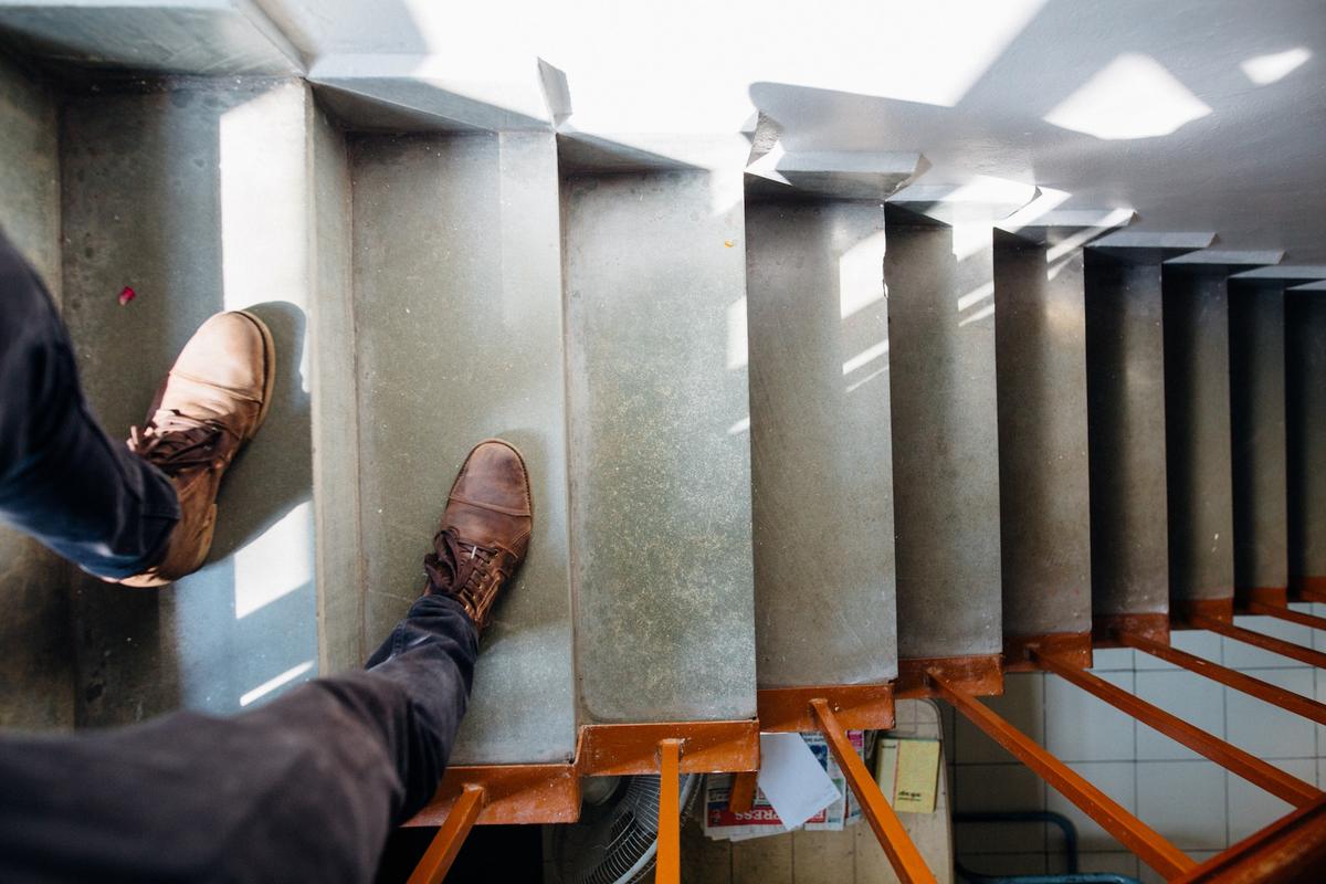 Overhead view of person's feet in brown laced shoes standing at top of concrete stairwell with orange metal railings, looking down multiple flights of stairs