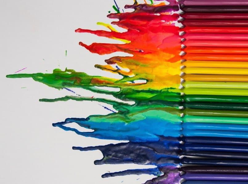 A row of colored pencils arranged vertically on the right side, with their wax tips melted and blown across a white canvas to the left in a vibrant, rainbow-colored splash pattern.
