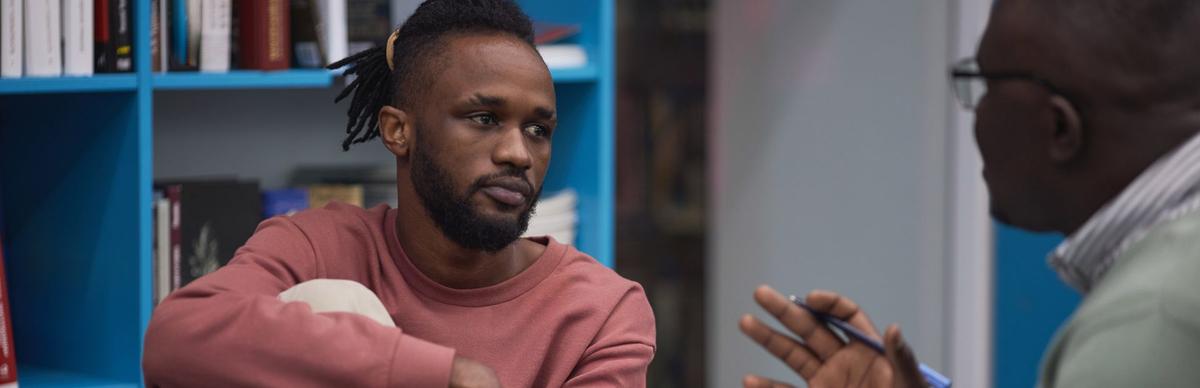 Close-up of a young Black man with a thoughtful expression looking toward an out-of-focus person who is gesturing with a pen.