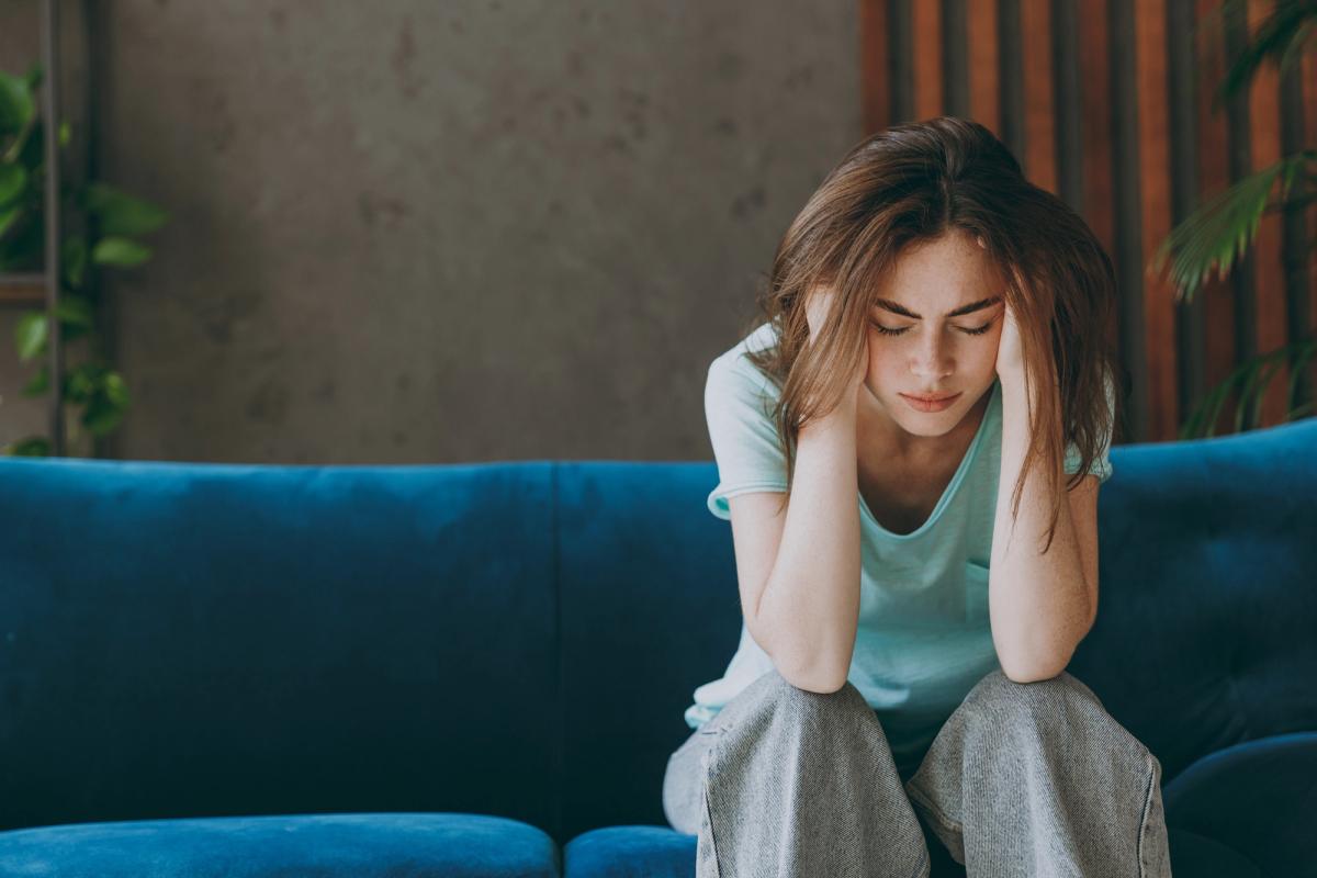 Young woman sitting on a couch with her head in her hands showing emotional distress, anxiety, or depression in a quiet home setting.