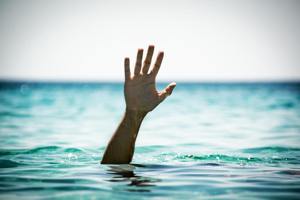 A person's arm and open hand reach upward out of turquoise ocean water. The person is submerged, and the horizon line is visible in the background under a bright, clear sky.