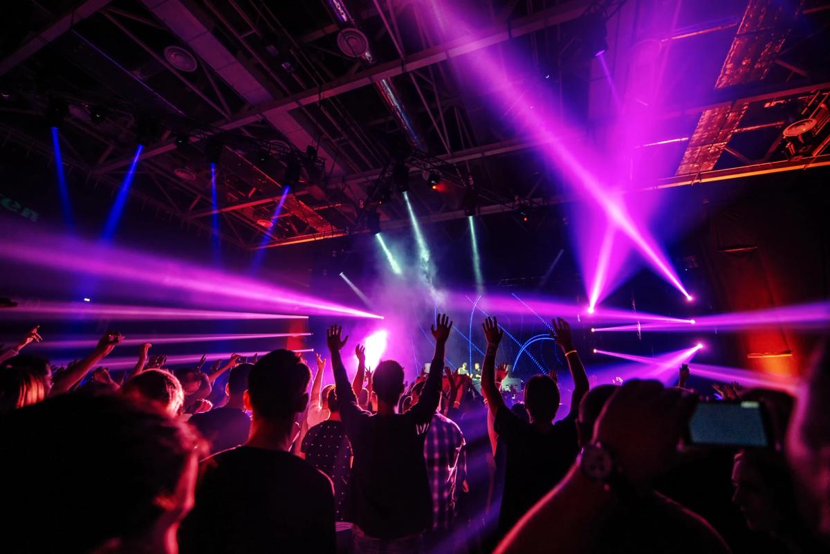 Crowd cheering with hands raised at an indoor concert or nightclub, illuminated by purple and pink laser lights and stage beams in a large venue.