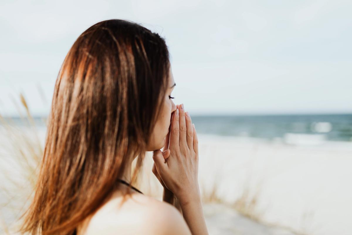 A woman with long auburn hair stands on a beach, her hands held up to her face in a prayer-like or thoughtful gesture while looking out at the ocean.