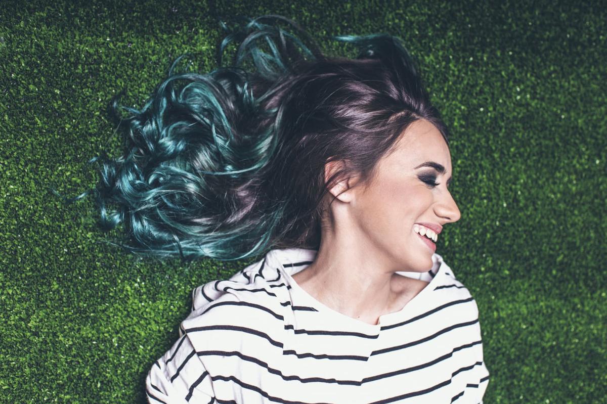 A smiling woman with flowing, dark and teal-colored hair lies on a patch of bright green grass.