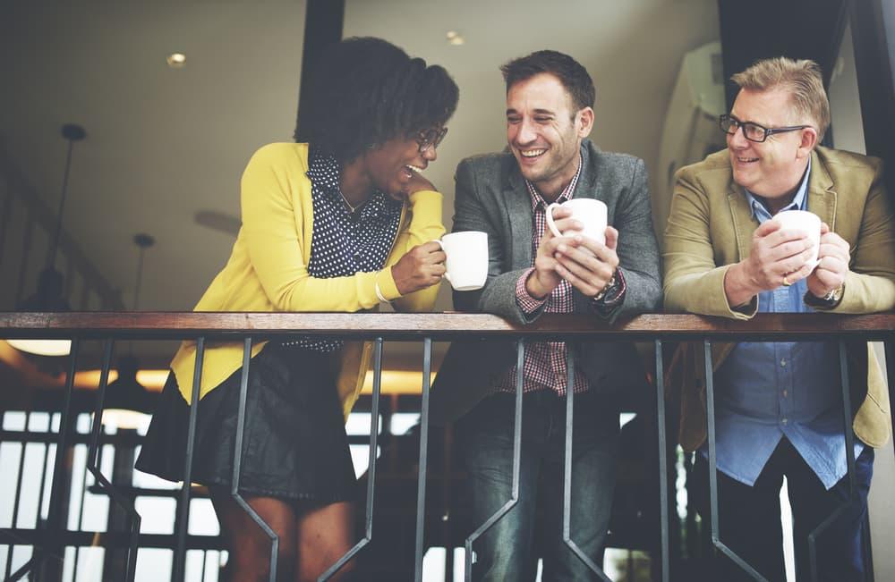 Three coworkers lean on a railing and laugh together while holding coffee mugs, representing supportive workplace connections and rebuilding professional life during recovery.
