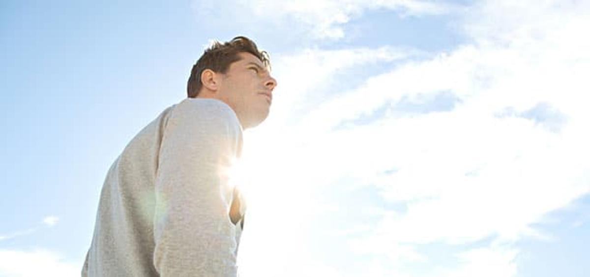 A young man in a grey sweater looking upward toward a bright, sun-filled sky with light wispy clouds.