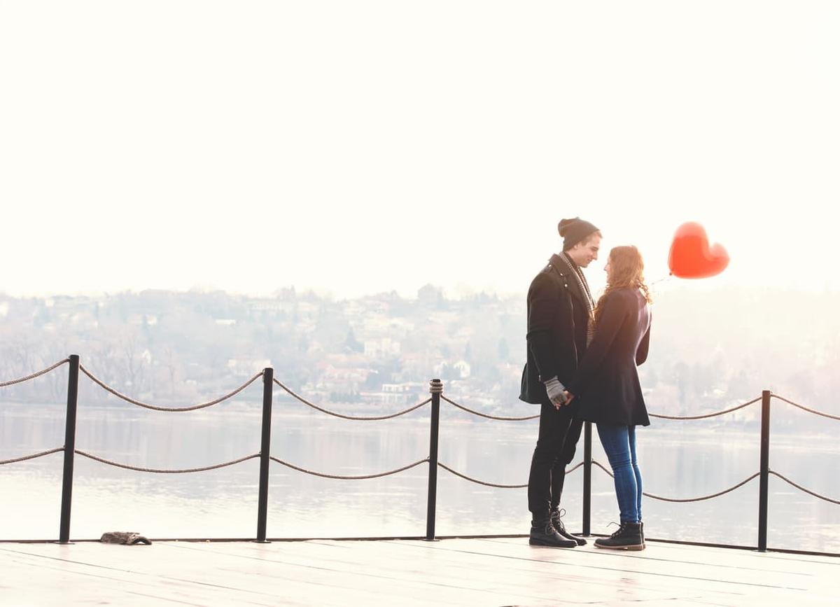 A couple standing on a bridge, looking into each other's eyes, while holding a heart-shaped balloon.