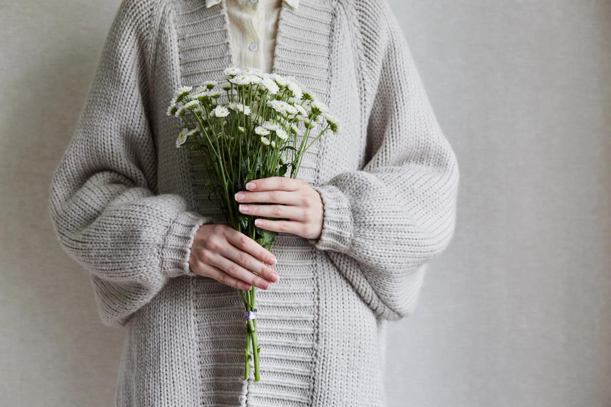 A person wearing a thick, grey oversized knit cardigan holding a bunch of small white daisies.