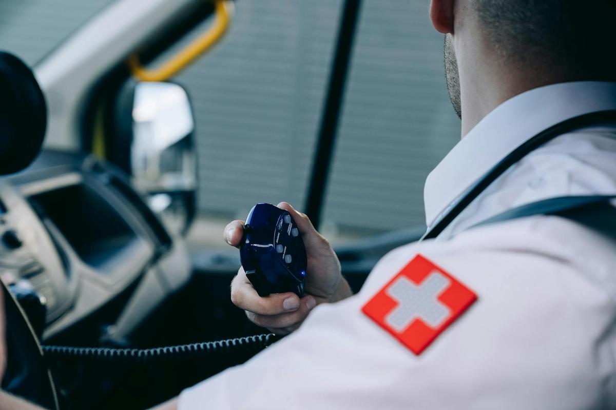Over-the-shoulder view of a paramedic in a white uniform with a red medical cross patch, holding a blue radio microphone inside the cabin of an ambulance.