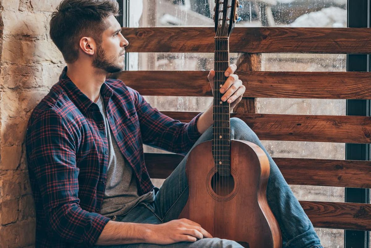 Man sitting by a window holding an acoustic guitar and looking outside on a rainy day.