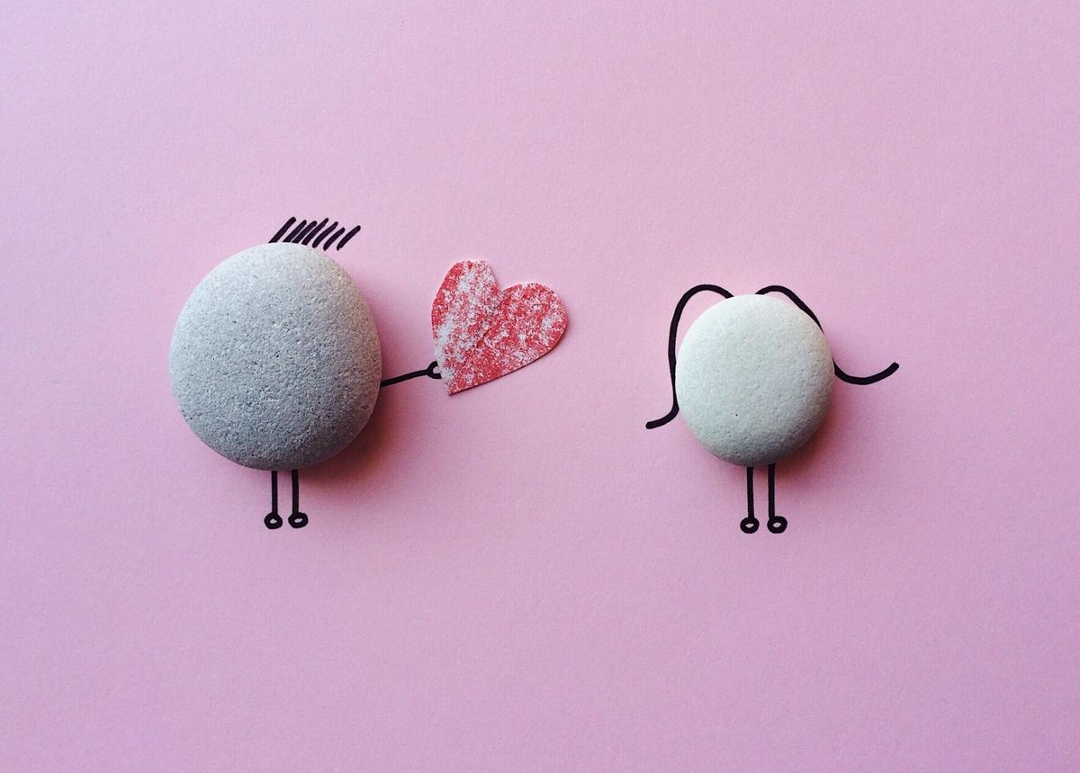 A creative illustration on a pink background featuring two smooth gray stones transformed into minimalist figures with hand-drawn ink lines.