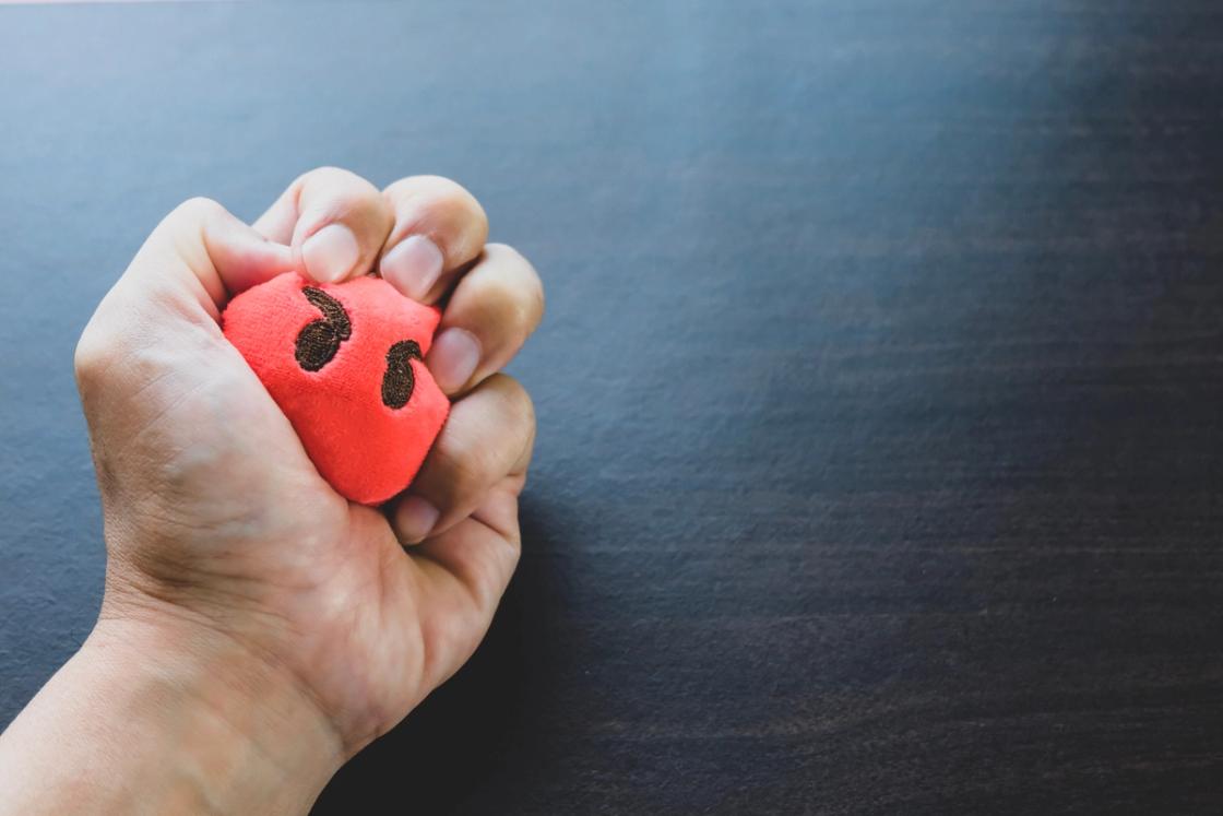 A close-up shot of a person’s hand tightly squeezing a small, red plush heart against a dark, textured wooden surface.