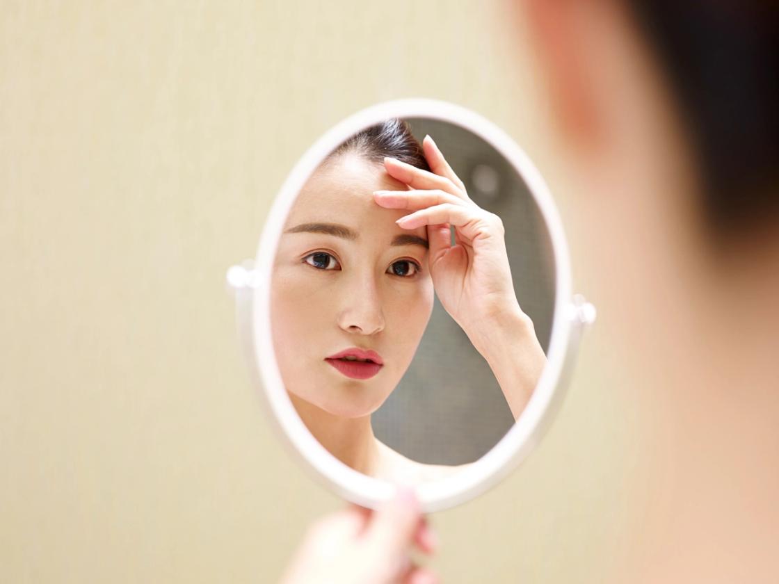 A woman looks closely at her reflection in a small, round vanity mirror while touching her forehead.