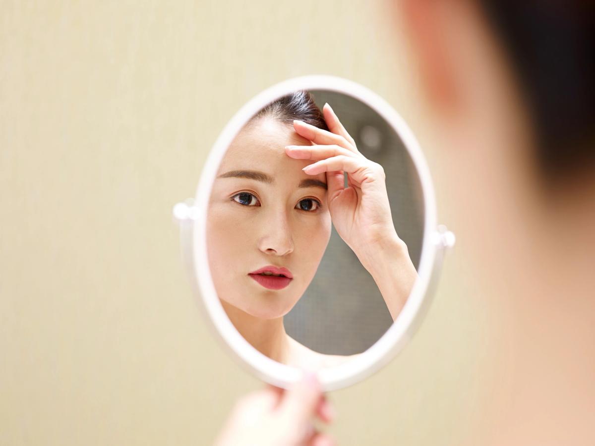 A woman looks closely at her reflection in a small, round vanity mirror while touching her forehead.