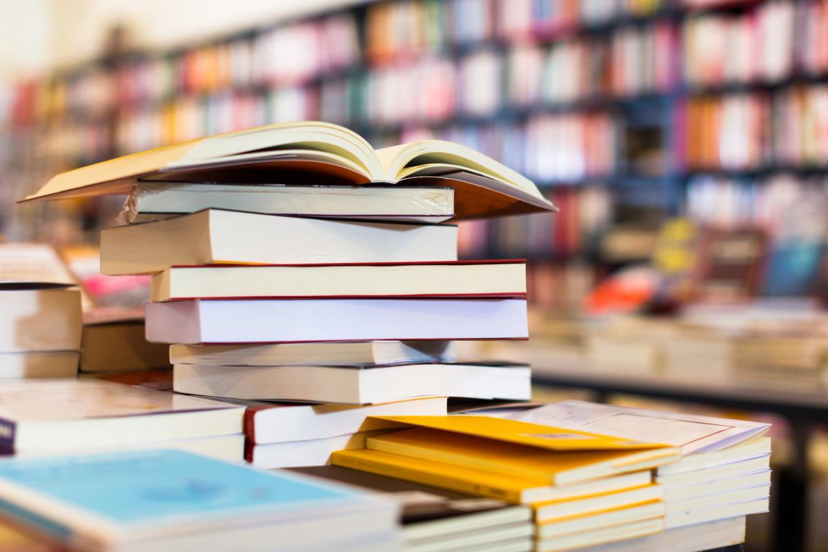 A tall, slightly messy stack of paperback and hardcover books on a table, with an open book resting on top, set against the blurred background of a bookstore with floor-to-ceiling shelves.