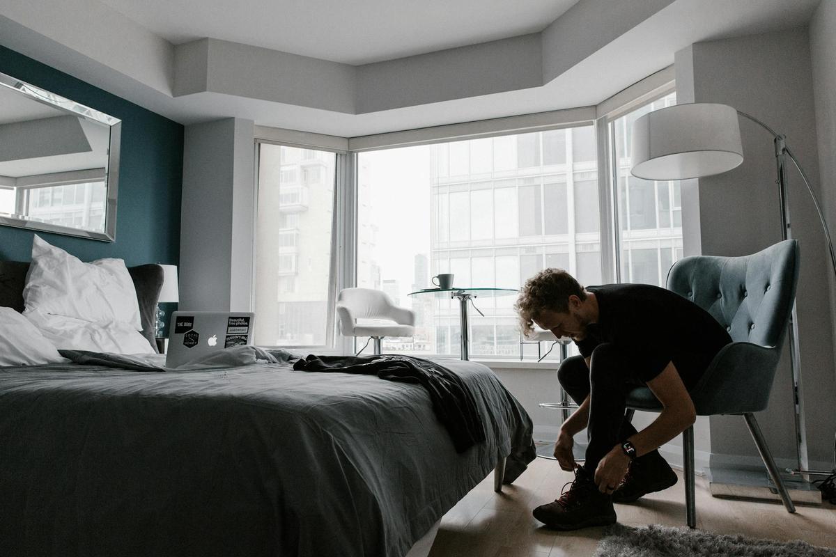 A man with curly hair sits in a modern, light-filled bedroom, leaning over to tie his hiking boots.