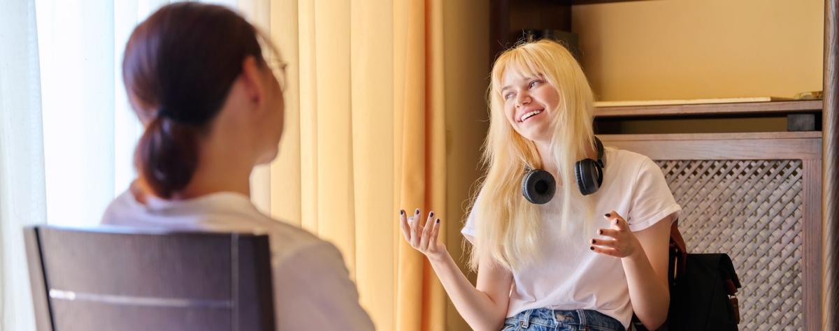 A young blonde woman with headphones around her neck smiles and gestures with her hands while talking to a person sitting across from her in a brightly lit room.