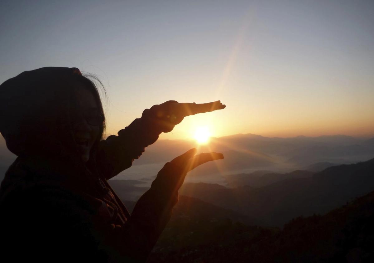 A silhouette of a person standing on a mountain at sunrise, holding their hands out to "frame" the sun between their palms.