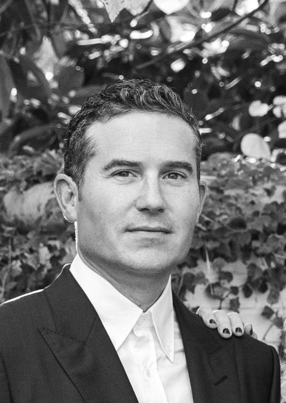 A black and white portrait of a man with short, wavy hair wearing a dark suit jacket and a white collared shirt.