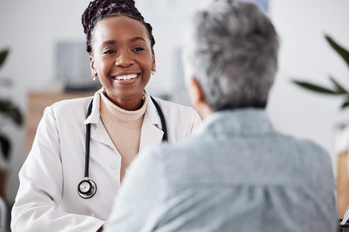 Smiling female healthcare provider wearing a white coat and stethoscope speaks with an older patient during a medical consultation.