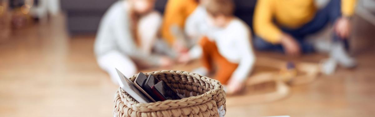 Woven basket filled with smartphones in the foreground while a family spends time together in the background, illustrating digital burnout and the choice to unplug from screens.