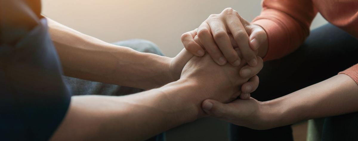 A close-up of two people holding hands in a supportive gesture. One person’s hands are gently clasped over the other’s, conveying comfort and empathy in a soft-focus setting.