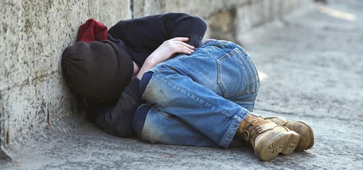 A homeless person in a fetal position, wearing a dark jacket, black beanie, jeans, and work boots, sleeping on the pavement next to a concrete wall.