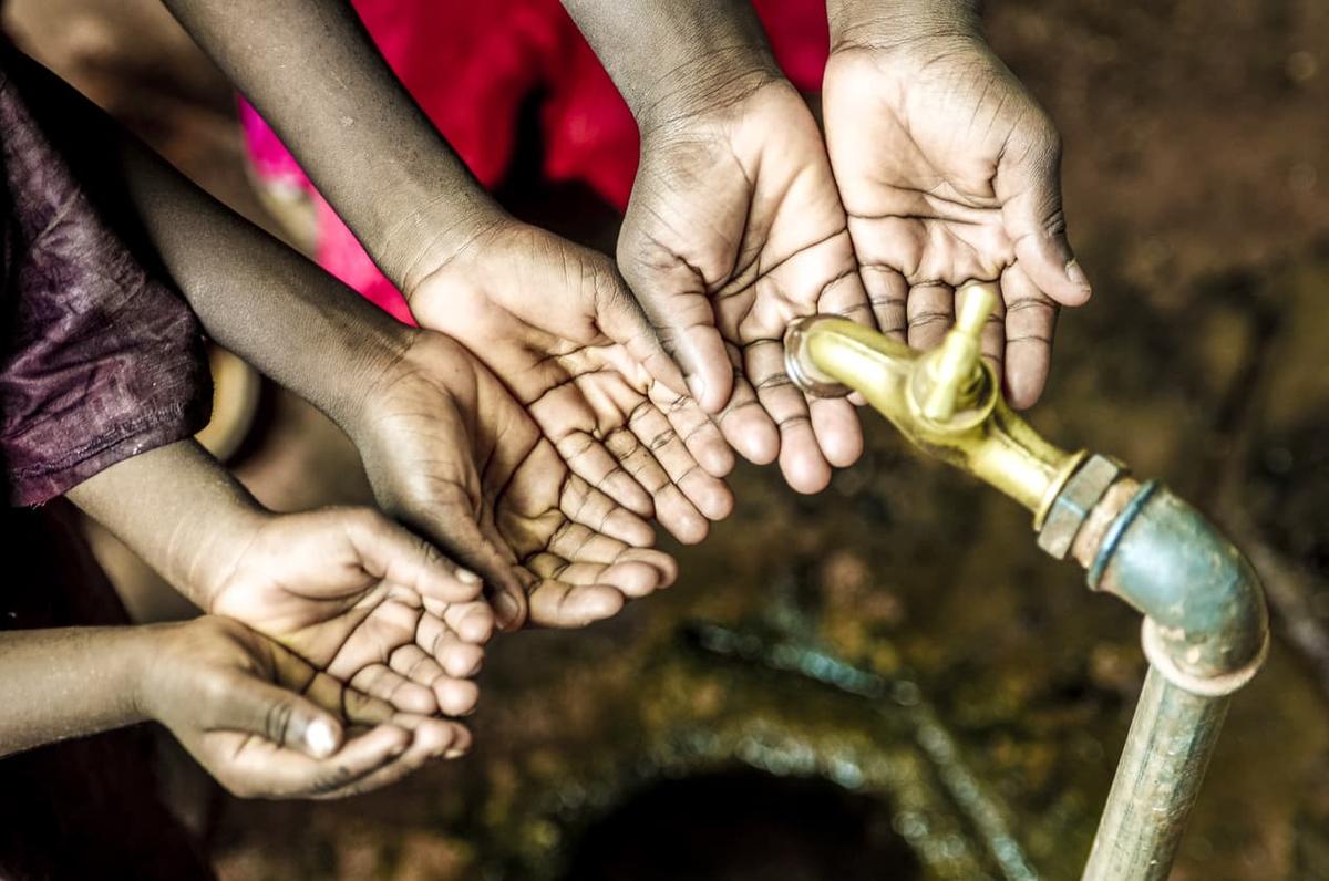 Several pairs of hands are cupped together beneath a brass water spigot.