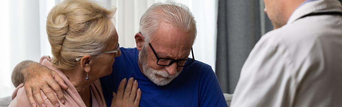 An elderly woman comforting a distressed elderly man during a medical consultation.