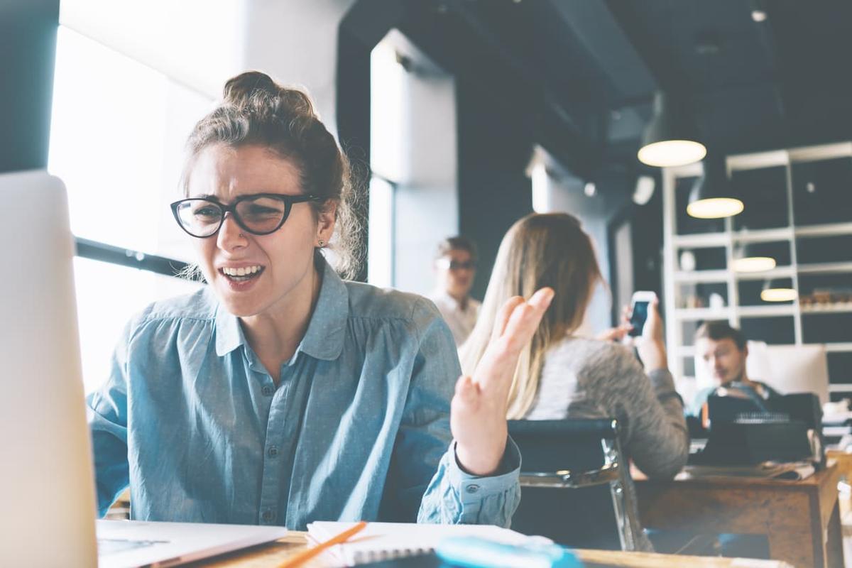 A young woman with glasses and a bun looks frustrated and confused while working on a laptop in a busy modern office.