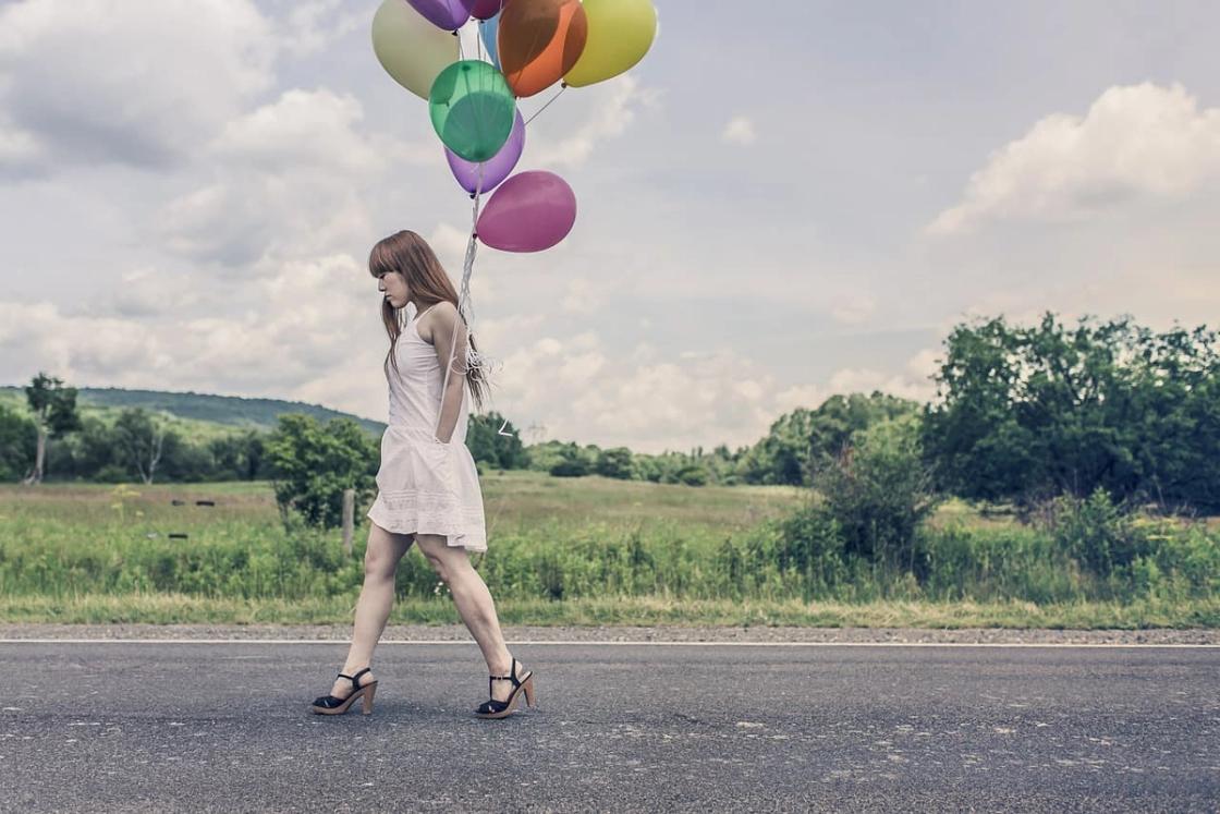 A woman in a white dress and high heels, carrying colorful balloons, and walking down a street.