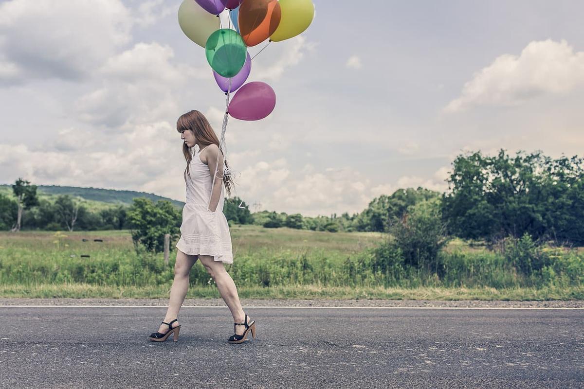 A woman in a white dress and high heels, carrying colorful balloons, and walking down a street.