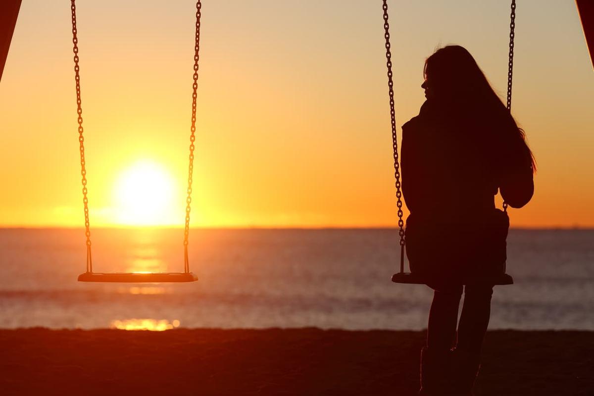 Silhouette of a woman sitting on a beach swing at sunset, looking out toward an empty swing beside her and the orange sun reflecting over the ocean.