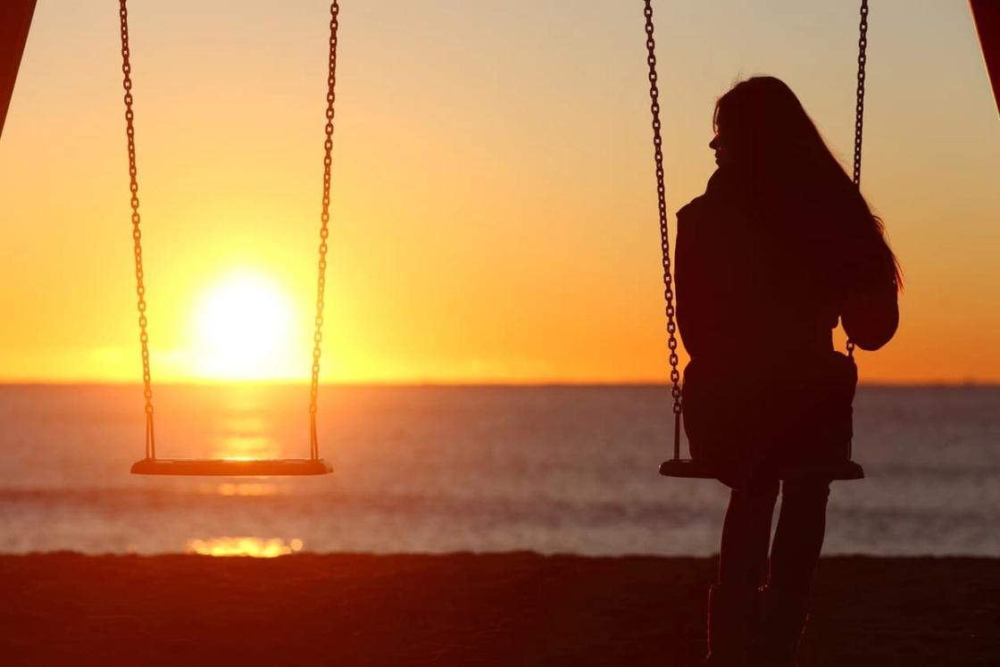 Silhouette of a woman sitting on a beach swing at sunset, looking out toward an empty swing beside her and the orange sun reflecting over the ocean.