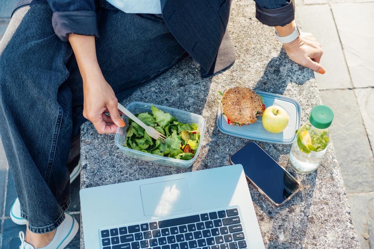 Person sitting outdoors with a healthy lunch of salad, sandwich, apple, and infused water beside a laptop and phone, symbolizing the difference between habit vs addiction and the role of intentional daily choices in recovery.