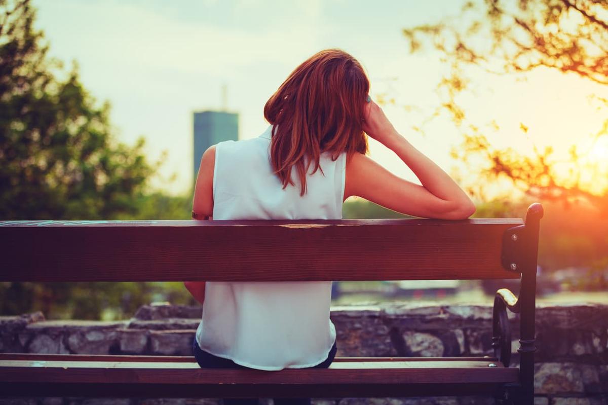 Woman sitting alone on a park bench at sunset, facing away and looking out over a city skyline, conveying solitude and reflection.