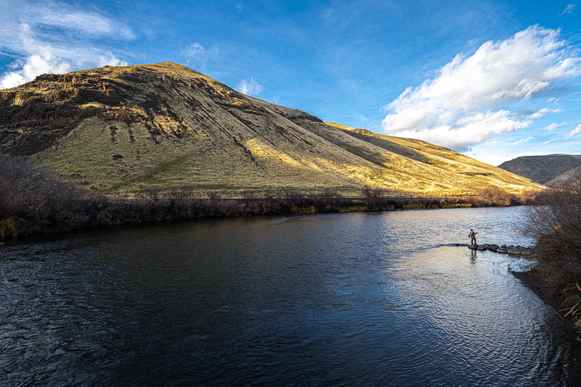 A wide river flowing past a large, sun-drenched grassy hill under a bright sky with scattered clouds, with a lone person standing near the water's edge