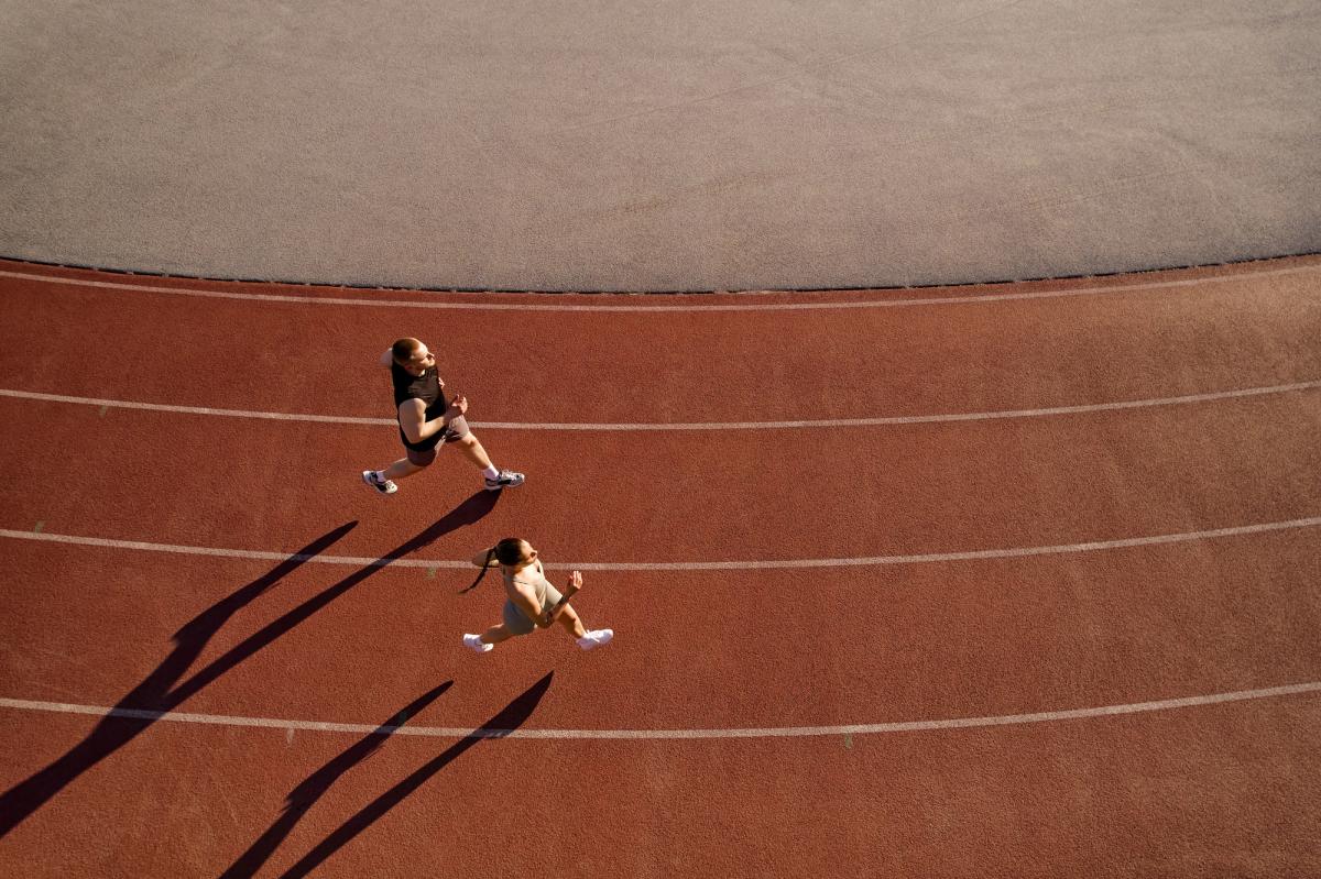 Two runners jogging on an outdoor track in the sunlight, captured from above with long shadows on the red track surface.