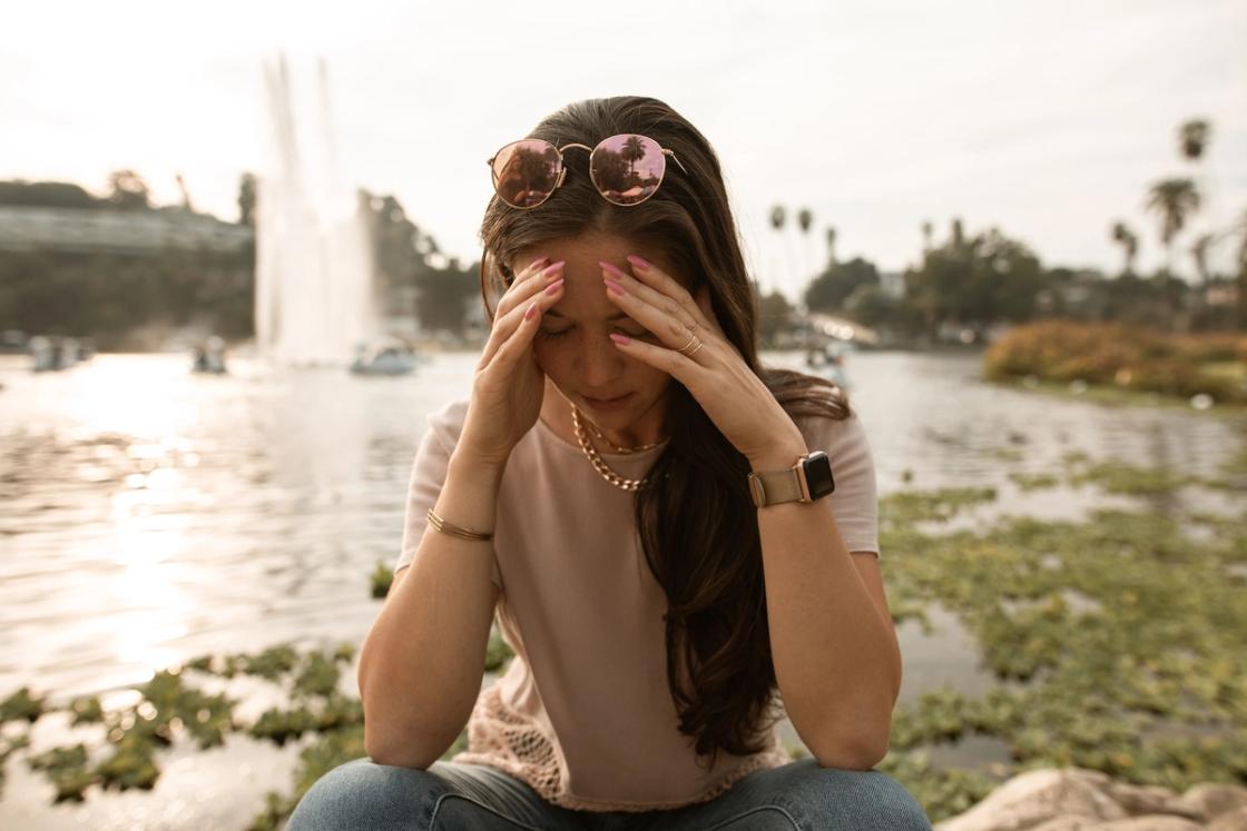 Woman sitting by a lake with hands on her temples, appearing stressed or overwhelmed, with a fountain and trees in the background.