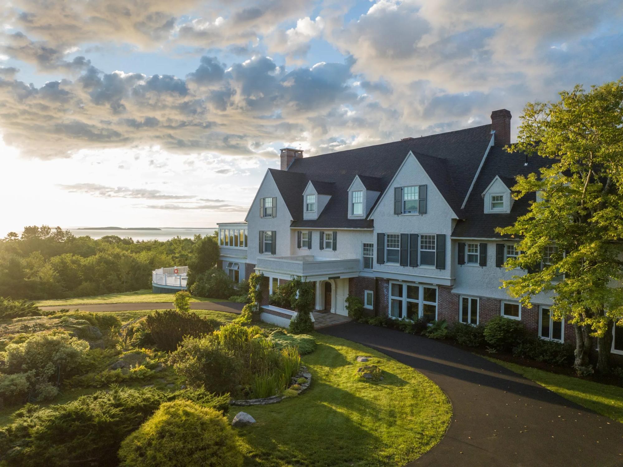 A wide aerial view of a grand, multi-story estate with light gray siding, dark shingles, and several gables.
