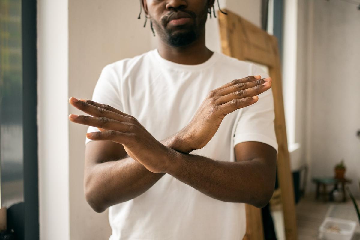 A close-up, waist-up shot of a Black man wearing a plain white t-shirt. He has his arms crossed in front of his chest in an "X" formation, with his palms facing outward and fingers extended.