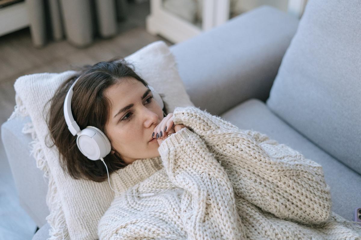 Woman lying on grey couch wearing white headphones and wrapped in cream knit blanket, appearing peaceful and relaxed