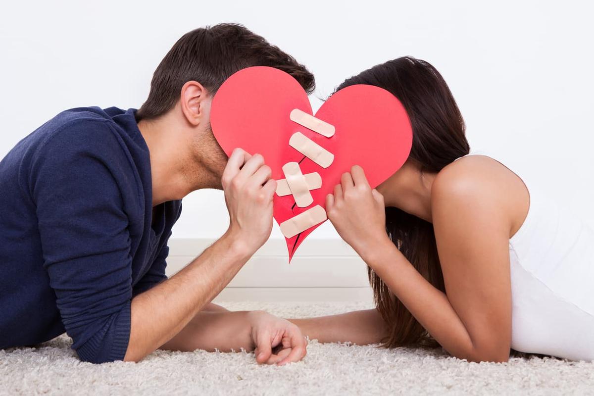 A man and a woman lying on a plush white rug, facing each other and hiding their faces behind a large red paper heart that has been mended with several bandages.